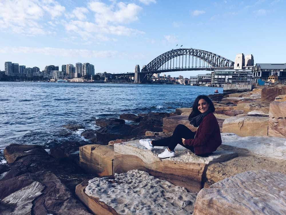 Estudiante en Australia chica morena sentada en unas rocas y detrás la ciudad de Sydney y sonriendo a cámara